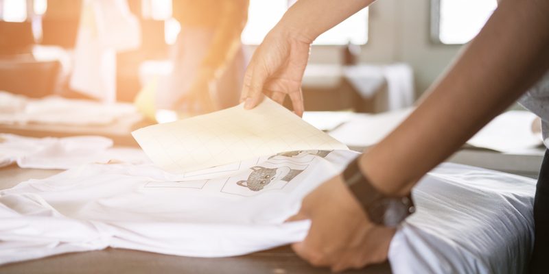 Young woman pull out paper from waterproof film on fabric at shop. worker working on manual screen printing on t-shirt. worker working on manual screen printing on t-shirt at her shop.